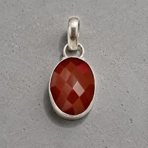May include: A pendant featuring a faceted, oval-shaped deep red stone set in a smooth silver-toned metal bezel. The pendant includes a sturdy, rounded silver-toned metal bail at the top, designed for attachment to a chain or cord necklace.