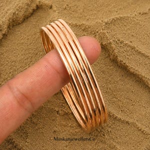Peut inclure: Un ensemble de cinq joncs fins, lisses et de couleur cuivre métallique avec une finition polie. Les bracelets circulaires sont empilés ensemble, présentant un design simple et sans ornement avec une surface réfléchissante.