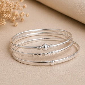Puede incluir: Un conjunto de tres brazaletes plateados. El brazalete superior presenta una esfera pulida central con detalles acanalados. El brazalete central muestra una fila de cinco pequeños corazones en relieve. El brazalete inferior incluye un corazón.