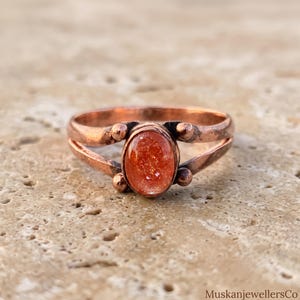 May include: A copper ring featuring an oval-shaped, glittery orange sunstone cabochon set in the center. The band has a split design with four small, round copper beads accenting the stone setting. The text MuskanjewellersCo is visible.