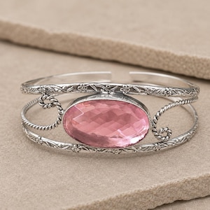Könnte beinhalten: Ein silbernes Armband mit einem ovalen, facettierten rosa Edelstein in der Mitte. Das Metallband weist an den Rändern komplizierte florale Gravuren und gedrehte Drahtdetails auf, die mit der zentralen Steinfassung verbunden sind.
