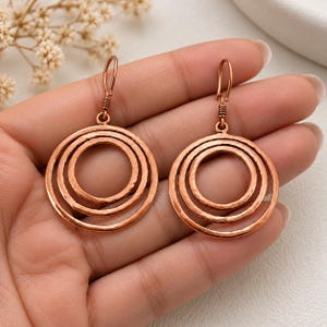 May include: A pair of copper-toned dangle earrings featuring three concentric circles with a hammered texture. Each earring is suspended from a simple hook wire, showcasing a metallic finish and a layered circular design that creates a distinct visual depth.
