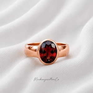 Peut inclure: Une bague en or rose ornée d'une grande pierre grenat rouge foncé de taille ovale, montée sur un anneau lisse et poli. La bague est centrée, et le texte MuskanjewellersCo est imprimé en police cursive noire sous le bijou.