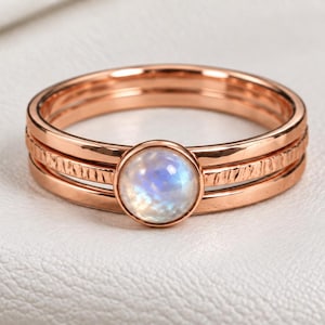 Op de afbeelding: Een roségouden ring met een centrale ronde maansteen cabochon met een blauwe iriserende glans. De ring bestaat uit drie gestapelde lagen, waarbij de buitenste twee banden een getextureerde, gehamerde afwerking hebben en de middelste band de steen vasthoudt.