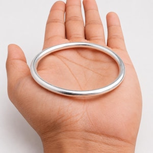 Op de afbeelding: Een gladde, gepolijste zilverkleurige armband met een eenvoudig, afgerond en doorlopend cirkelvormig ontwerp. Het metalen oppervlak is reflecterend en uniform, wat een strakke esthetiek laat zien zonder extra versieringen of patronen.