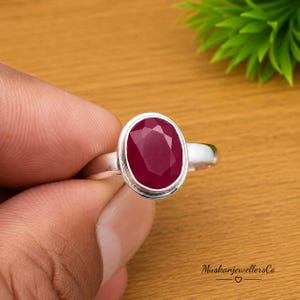 May include: A silver ring featuring a faceted oval-shaped deep red ruby gemstone set in a smooth metal bezel. The ring band is polished silver and the stone has a multi-faceted surface that reflects light, creating a vibrant appearance.