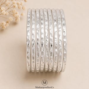 Peut inclure: Un ensemble de neuf bracelets rigides de couleur argentée disposés en pile verticale. Chaque bracelet présente une surface martelée et texturée qui reflète la lumière. Le nom de marque MuskanjewellersCo avec un logo M est imprimé en bas.
