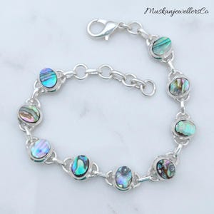May include: A silver chain bracelet featuring eight oval-shaped abalone shell inlays with iridescent blue, green, and purple tones. The bracelet includes a lobster claw clasp and a series of circular links. The text MuskanjewellersCo is visible.