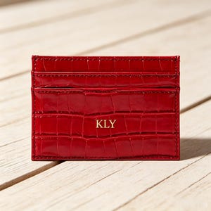 May include: A rectangular red card holder with a glossy crocodile skin texture. The front features the gold embossed letters KLY centred in the middle. The item includes multiple horizontal slots for organising cards and has visible stitching.