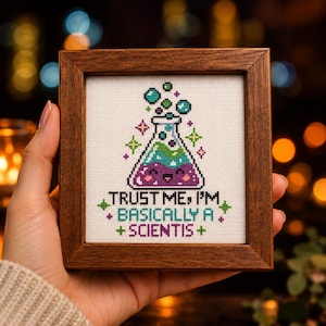 Könnte beinhalten: Ein quadratischer Holzrahmen mit einer Kreuzstichstickerei auf weißem Stoff. Das Motiv zeigt einen lächelnden violett-grünen Chemiekolben mit Blasen und den Text: TRUST ME, I'M BASICALLY A SCIENTIS. Die Stickerei verwendet leuchtende Garnfarben.