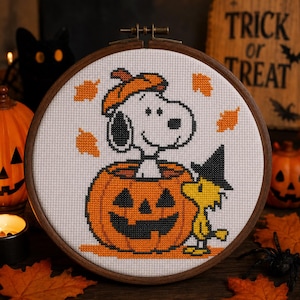 Puede incluir: Un bastidor de bordado circular de madera con un patrón de punto de cruz de un perro blanco con una boina naranja dentro de una calabaza, acompañado por un pequeño pájaro amarillo con un sombrero de bruja negro y hojas naranjas.