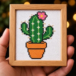 Può includere: Una cornice quadrata in legno contenente un ricamo a punto croce di un cactus verde con un fiore rosa in un vaso arancione. Il cactus presenta spine a punto croce bianco su uno sfondo di tessuto bianco con un motivo a griglia visibile.