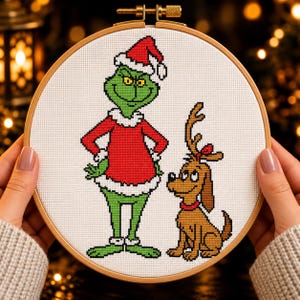 Puede incluir: Un bastidor de bordado circular de madera que contiene un diseño de punto de cruz sobre tela blanca. El diseño muestra a un personaje verde con traje y gorro de Papá Noel rojo, de pie junto a un perro marrón con una cornamenta y un collar rojo.