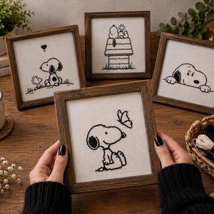 May include: Four square wooden frames containing black and white cross-stitch embroidery art featuring the character Snoopy. One frame shows Snoopy with a butterfly, another with Woodstock, one on a doghouse, and one peeking over a ledge.