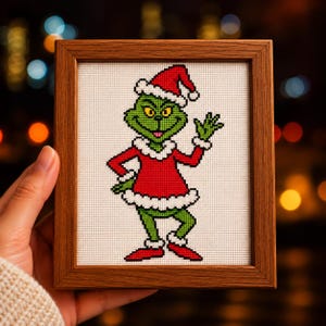 May include: A cross-stitch embroidery of a green creature with yellow eyes, wearing a red Santa suit with white fur trim and a matching hat, framed in a brown wooden square frame. The character is waving one hand and standing on white fabric.
