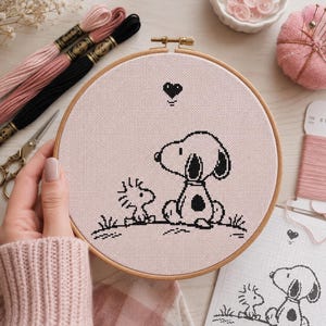 以下が含まれることがあります： 薄いピンク色の布が入った円形の木製刺繍枠。黒いクロスステッチで犬と小さな鳥が一緒に座っている様子が描かれています。キャラクターの上には小さな黒いハートが刺繍されており、横には対応する紙の図案が見えています。