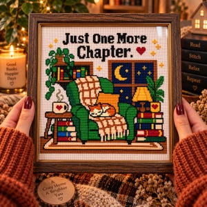 May include: A framed cross-stitch embroidery featuring the text Just One More Chapter with a red heart. The design depicts a green armchair with a plaid blanket, a sleeping cat, stacks of books, a houseplant, and a window showing a crescent moon.