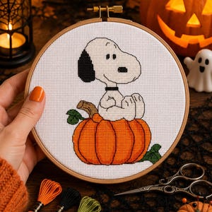 May include: A circular wooden embroidery hoop containing a cross-stitch design of a white dog with a black ear and collar sitting inside a large orange pumpkin with green leaves, set against a white fabric background with a grid pattern.