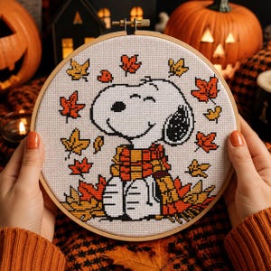 May include: A circular wooden embroidery hoop containing a cross-stitch pattern of a white cartoon dog wearing a red and orange plaid scarf. The dog is surrounded by scattered autumn leaves in shades of yellow, orange, and red on a white fabric.