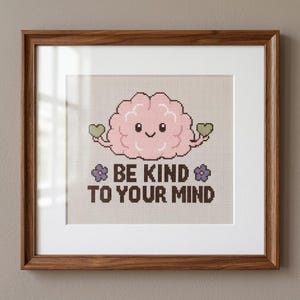 Puede incluir: Una obra de punto de cruz enmarcada que muestra un cerebro de dibujos animados de color rosa claro con una cara sonriente, sosteniendo dos pequeños corazones verdes. Debajo, el texto BE KIND TO YOUR MIND en letras marrón oscuro y dos flores moradas.