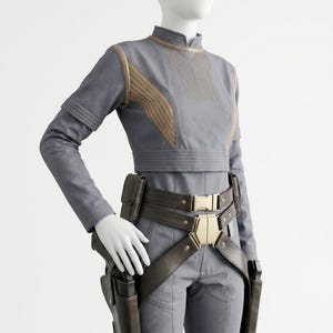 May include: A grey long-sleeved top with tan ribbed accents on the chest and shoulders, paired with matching grey trousers. A dark brown utility belt with a gold-toned buckle and two attached holsters is worn around the waist over the clothing.