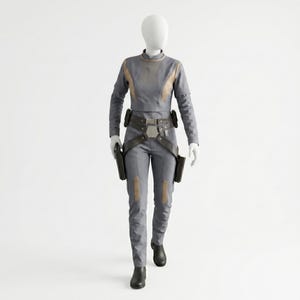 May include: A grey long-sleeved top and matching trousers set on a mannequin. The outfit features tan accents on the chest and legs, a dark brown utility belt with a central buckle, and two dark brown holsters attached to the sides of the belt.