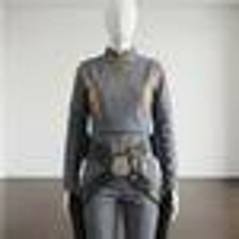 May include: A long-sleeved grey top featuring a high neckline and a textured, patterned fabric. The garment includes a dark, wide belt cinched at the waist with a prominent central buckle, paired with matching grey trousers in a solid, neutral tone.