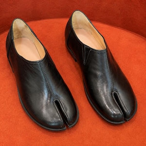 May include: A pair of black leather shoes featuring a distinct split-toe design. The shoes have a smooth finish, a low-cut topline, and a central seam running from the split toe to the opening. The interior lining is a light beige color.