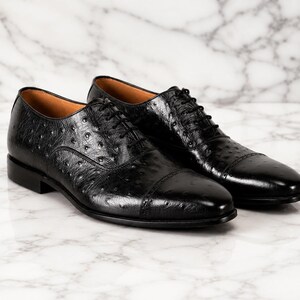 May include: A pair of black ostrich leather oxford shoes featuring a cap toe design, a lace-up closure with thin black laces, and a smooth tan interior lining. The textured leather surface displays the characteristic quill pattern.