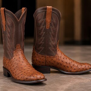 May include: A pair of western-style boots featuring a tan, textured ostrich leather foot with a distinct quill pattern. The tall, dark brown leather shafts are adorned with intricate tonal stitching and include matching tan leather pull straps.