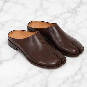 May include: A pair of dark brown leather mules featuring a distinctive split-toe design. These slip-on shoes have a smooth finish, a low heel, and a tan interior lining, showcasing a clean silhouette with a prominent seam running down the center.