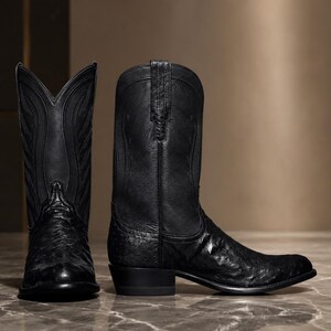 Puede incluir: Un par de botas de estilo vaquero en color negro con un patrón texturizado en el empeine y la punta, una caña de cuero liso con detalles de costuras tonales, punta afilada y un tacón apilado que completa su silueta clásica.