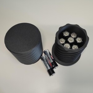 Könnte beinhalten: Ein schwarzer, zylindrischer Batterie-Aufbewahrungsbehälter mit strukturierter Oberfläche und abnehmbarem Deckel. Der offene Behälter enthält sieben AA-Batterien, während zwei weitere AA-Batterien horizontal daneben liegen.