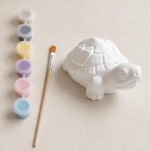 May include: A white ceramic turtle figurine with a textured shell, accompanied by a wooden paintbrush and a strip of six small paint pots containing beige, dark grey, yellow, purple, pink, and light blue paint colours for creative projects.