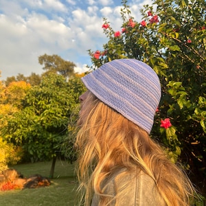 May include: A crochet bucket hat featuring horizontal stripes in alternating shades of light blue and soft lavender. The hat has a rounded crown and a short, downward-sloping brim, constructed with a dense, uniform stitch pattern throughout.