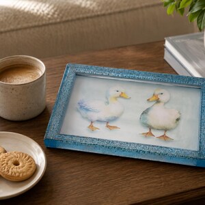 May include: A rectangular picture frame with a textured, glittery blue border containing an illustration of two white ducks with yellow beaks and orange feet standing on a light blue background, positioned next to a ceramic mug and a small plate.
