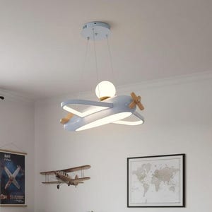 May include: A light blue aeroplane-shaped ceiling pendant light fixture featuring a central white spherical globe, wooden propeller accents, and integrated LED lighting panels along the wings, suspended from the ceiling by thin metal wires.
