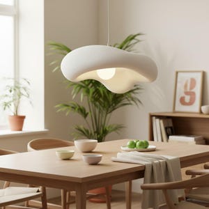 May include: A white pendant light with an organic, cloud-like shape hangs from a thin cord. The fixture features a textured matte surface and a central spherical bulb that emits a soft glow, suspended above a wooden dining table in a room.