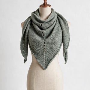 Könnte beinhalten: Ein dreieckiger Strickschal in Salbeigrün mit gerippter Textur und einer zentralen vertikalen Reihe von Ösendetails. Der Schal ist um eine Schaufensterpuppe drapiert und zeigt seine spitze Form sowie das gleichmäßige Strickmuster im gesamten Stoff.