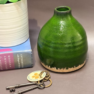 May include: A small, bulbous ceramic vase with a glossy, deep green glaze and a textured, unglazed beige base. Beside it lie two antique-style metal skeleton keys attached to a round tag that reads "GENUINE FOUR LEAF CLOVER GOOD LUCK CHARM."