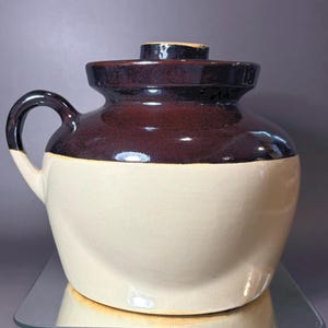 May include: A ceramic crock with a rounded body and a single side handle. The upper portion and lid are finished in a glossy dark brown glaze, while the lower portion features a smooth, matte cream-coloured finish. The lid has a small knob handle.