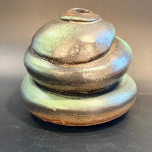 May include: A hand-built ceramic vessel with a stacked, rounded form. The piece features a dark, mottled glaze with prominent patches of vibrant green and earthy brown tones. It has a small, circular opening at the top and a smooth, glossy finish.