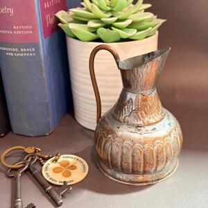 May include: A small, weathered copper-toned metal pitcher with a curved handle and embossed decorative patterns. Beside it lie two antique-style iron skeleton keys attached to a round wooden keychain featuring a four-leaf clover graphic and text.