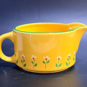 Puede incluir: Una salsera de cerámica de color amarillo brillante con un asa y un pico vertedor. El exterior está decorado con un patrón repetitivo de pequeñas flores blancas con tallos y hojas verdes, y el borde está acentuado con una fina línea verde.