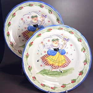 May include: Two round ceramic plates with scalloped blue edges. The border features a green lattice pattern with pink rosebuds. The center depicts a sheep dressed in a white blouse, black corset, blue sleeves, and a yellow and red plaid skirt.