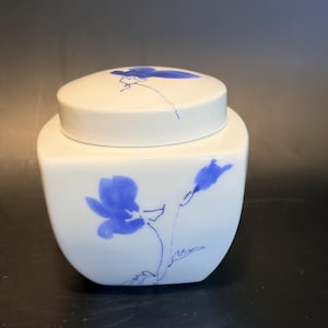 Könnte beinhalten: Ein weißes Keramikgefäß mit quadratischem Boden und passendem Deckel. Das Gefäß ist mit einem handgemalten blauen Blumenmuster mit feinen Pinselstrichen auf der Vorderseite und einem einzelnen blauen Blumenmotiv auf dem Deckel verziert.