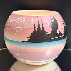Op de afbeelding: Een ronde keramische pot met een roze en crèmekleurige achtergrond met kleurverloop en een geschilderd zwart silhouet van bomen en een bergketen. Een horizontale turquoise band omcirkelt het onderste gedeelte en een witte handtekening is zichtbaar.