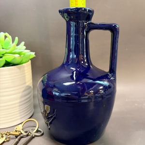 May include: A glossy dark blue ceramic pitcher with a single handle and a narrow neck. A yellow measuring tape is positioned at the top opening, showing a height measurement of approximately 8 inches. The pitcher has a smooth, reflective surface.