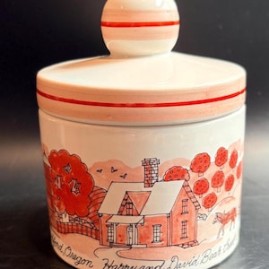 Puede incluir: Un tarro de cerámica blanca con tapa que presenta una franja roja pintada. El cuerpo muestra un dibujo lineal en rojo de una casa, árboles y un carruaje de caballos, con el texto "Medford, Oregon Harry and David Bear Creek" en la base.