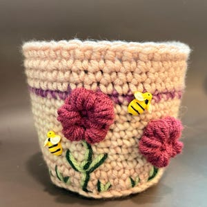 May include: A cylindrical crochet plant pot cover in light beige yarn featuring a horizontal purple stripe. The exterior is decorated with two dark pink crocheted flowers, green embroidered stems, and two small yellow and black bee buttons.